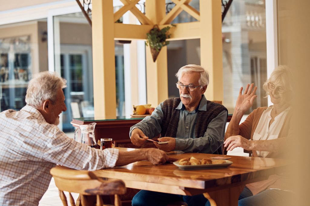 three residents talk and play a card game around a wooden table that also contains a pitcher of iced tea and a plate of patries