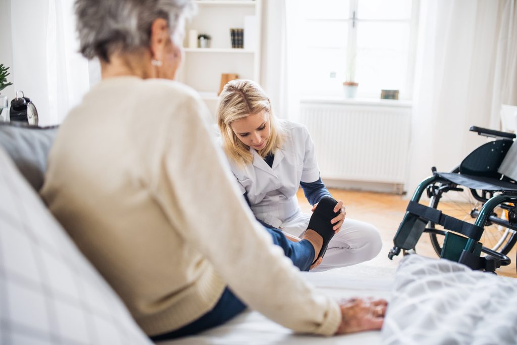 a caretaker helps a resident with shoes. the setting is within their home bedroom space, and a wheelchair awaits to the side.
