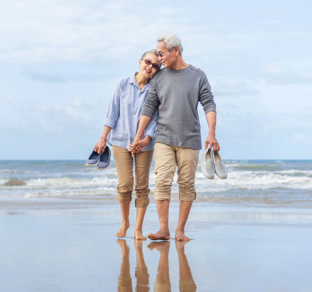 a couple hold hands and lean into each other, their feet bare on the wet send. rolling ocean waves and a partly cloudy sky are in the background