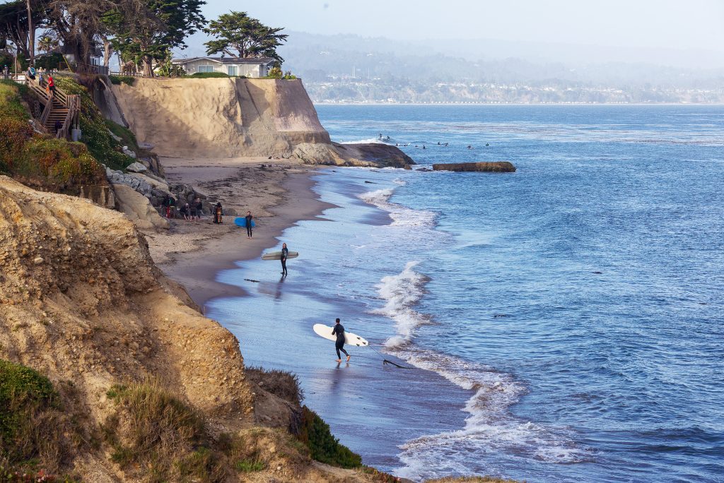 a view of the surf meeting the coastline and cliffs, where surfers are gathered in a cove. the coastline continues in the background, shrouded in mist