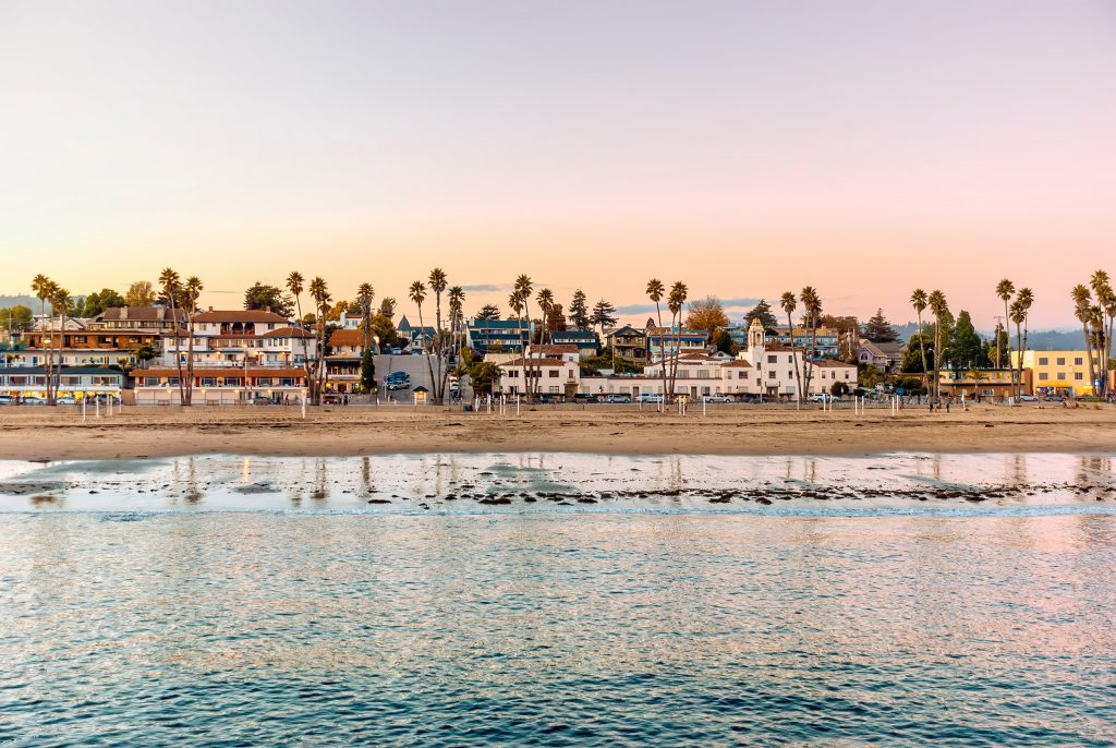 a view of the coastline with colorful buildings and palm trees across the middle, calm sea in the foreground, and a near-sunset sky in the background