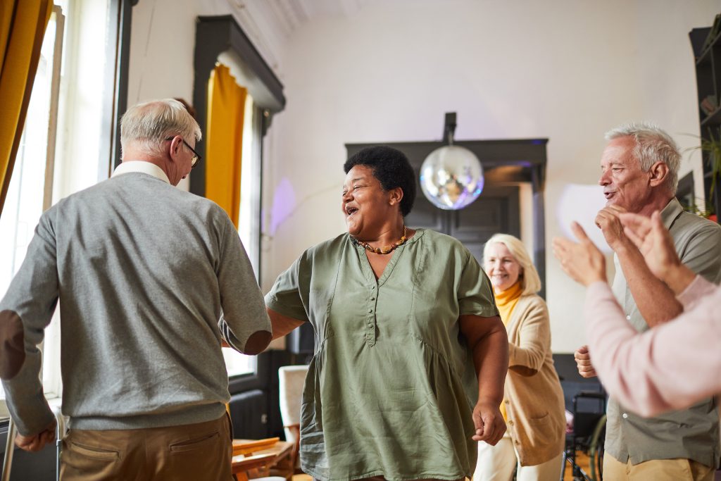 two residents in the foreground are dancing and smiling, while others around them watch and applaud, and a disco ball sparkles in the background