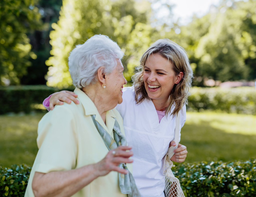a resident and younger person with her arm around the resident share a laugh and a smile while walking outdoors with grass, hedges and trees in the background