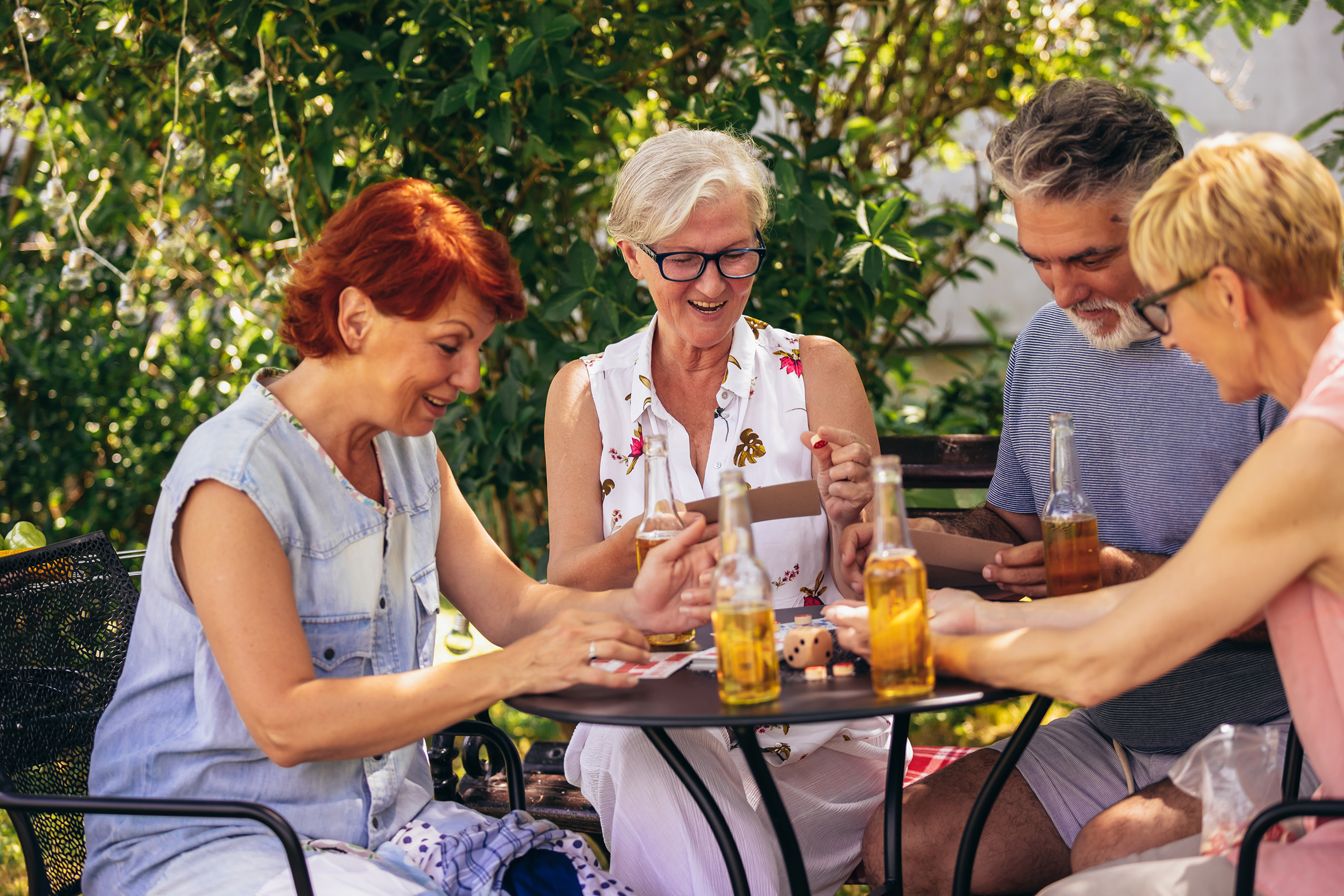 a group of residents share a game and beverages around a table outdoors, green hedges and dappled light fill the background