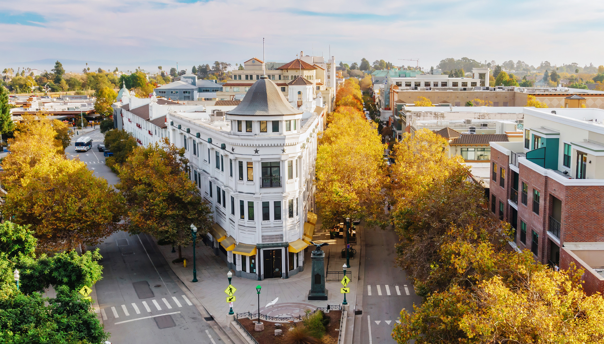 an aerial shot of a white wedge-shaped building within a narrow angled-intersection. trees with mostly yellow leaves line the streets