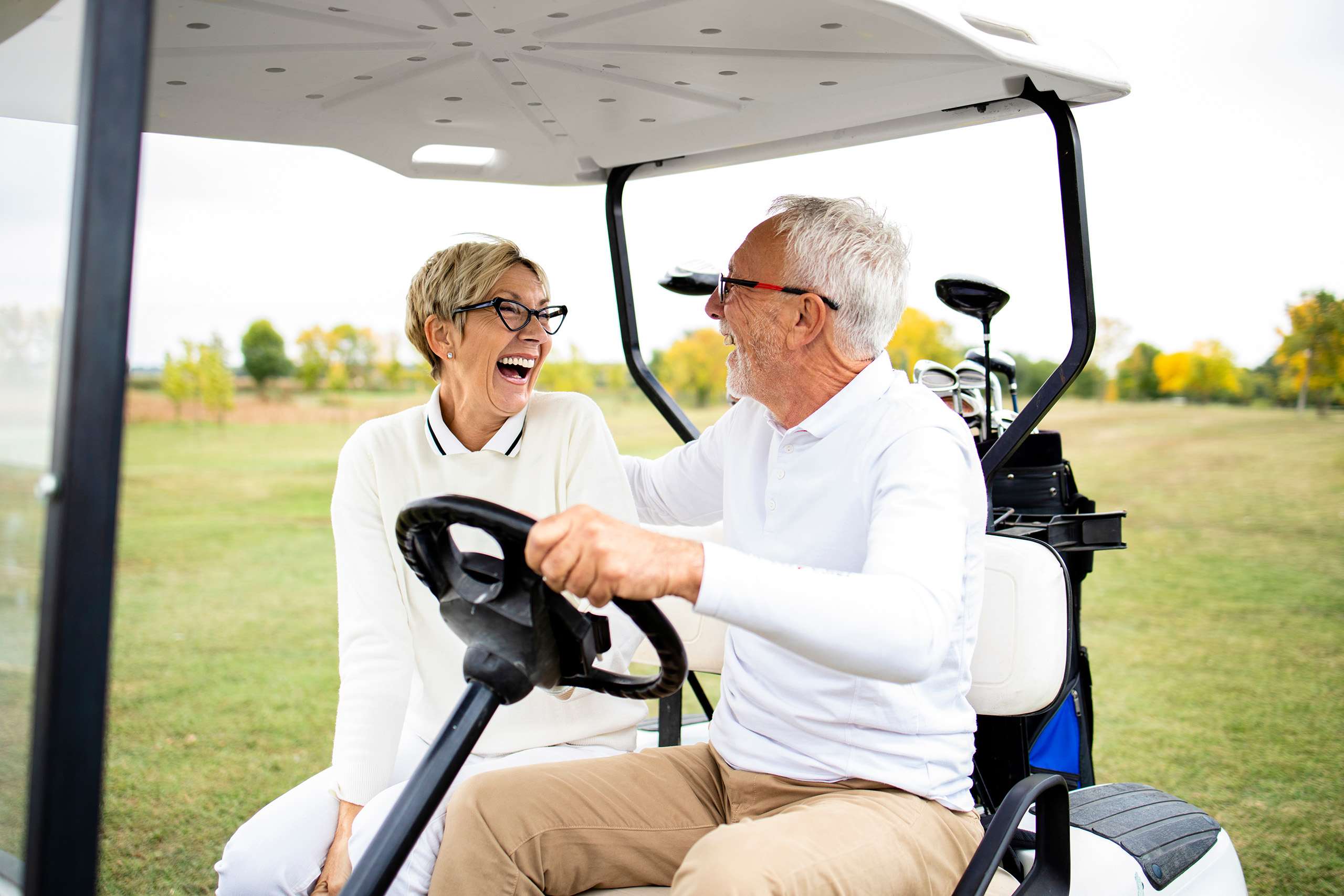a resident couple seated in a golf cart on a green lawn, looking towards each other and laughing